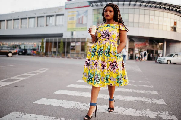 Cute small height african american girl with dreadlocks, wear at coloured yellow dress, walking at crosswalk against trade center with cup of coffee at hand.