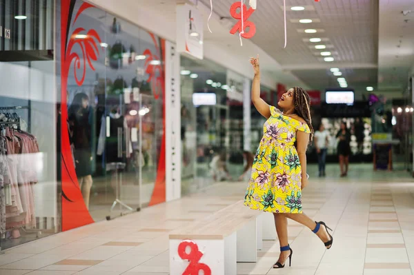 Cute small height african american girl with dreadlocks, wear at coloured yellow dress, showing finger on discount sign at mall.