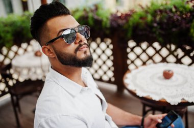 Şık uzun Arap adam modelinde beyaz gömlek, kot pantolon ve güneş gözlüğü şehir Caddesi'nde poz verdi. Açık kafe oturan ve cep telefonu arıyorum sakal çekici Arap adam.