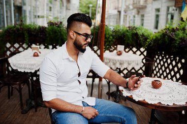 Şık uzun Arap adam modelinde beyaz gömlek, kot pantolon ve güneş gözlüğü şehir Caddesi'nde poz verdi. Açık kafe oturan ve cep telefonu arıyorum sakal çekici Arap adam.
