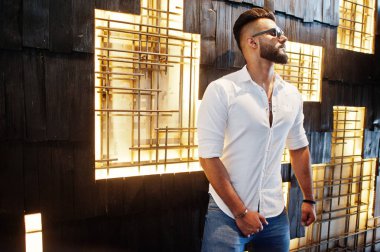 Beyaz gömlek, kot pantolon ve güneş gözlükleri şık uzun Arap adam modelinde ışık duvara kapalı poz verdi. Sakal çekici Arap adam.