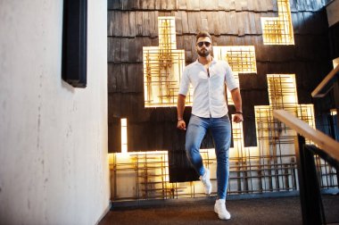 Beyaz gömlek, kot pantolon ve güneş gözlükleri şık uzun Arap adam modelinde ışık duvara kapalı poz verdi. Sakal çekici Arap adam.