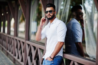 Şık uzun Arap adam modelinde beyaz gömlek, kot pantolon ve güneş gözlüğü şehir Caddesi'nde poz verdi. Sakal çekici Arap adam üstünde hareket eden telefon konuşma.