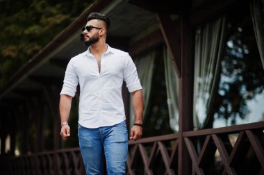 Şık uzun Arap adam modelinde beyaz gömlek, kot pantolon ve şehir Caddesi'nde yürürken güneş gözlüğü. Sakal çekici Arap adam.