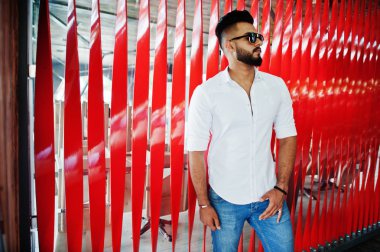 Şık uzun Arap adam modelinde beyaz gömlek, kot pantolon ve güneş gözlüğü şehir Caddesi'nde poz verdi. Sakal çekici Arap adam kırmızı duvara.