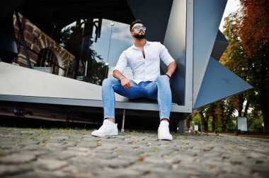 Şık uzun Arap adam modelinde beyaz gömlek, kot pantolon ve şehir Caddesi'nde oturan güneş gözlüğü. Sakal çekici Arap adam.