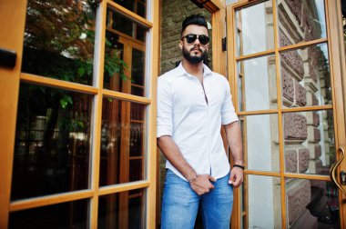 Şık uzun Arap adam modelinde beyaz gömlek, kot pantolon ve güneş gözlüğü şehir Caddesi'nde poz verdi. Sakal çekici Arap adam karşı giriş kapısı Restaurant.