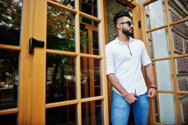 Şık uzun Arap adam modelinde beyaz gömlek, kot pantolon ve güneş gözlüğü şehir Caddesi'nde poz verdi. Sakal çekici Arap adam karşı giriş kapısı Restaurant.