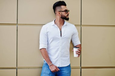 Şık uzun Arap adam modelinde beyaz gömlek, kot pantolon ve güneş gözlüğü şehir Caddesi'nde poz verdi. Kahve altın duvara sakal çekici Arap adamla.