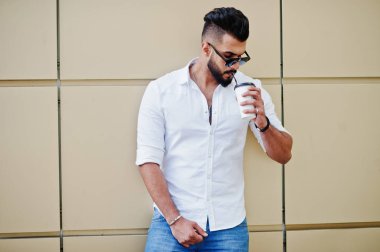 Şık uzun Arap adam modelinde beyaz gömlek, kot pantolon ve güneş gözlüğü şehir Caddesi'nde poz verdi. Bardak kahve altın duvara sakal çekici Arap adamla.