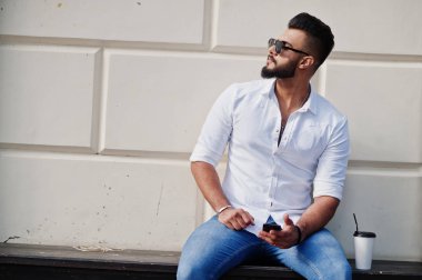 Şık uzun Arap adam modelinde beyaz gömlek, kot pantolon ve güneş gözlüğü şehir ve tutun cep telefonu eldeki Caddesi'nde poz verdi. Arap adam oturmuş ve dinlenme kahve ile sakal.
