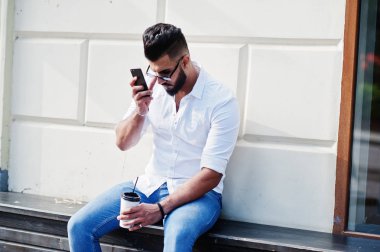 Şık uzun Arap adam modelinde beyaz gömlek, kot pantolon ve güneş gözlüğü şehir ve tutun cep telefonu eldeki Caddesi'nde poz verdi. Arap adam oturmuş ve dinlenme kahve ile sakal.