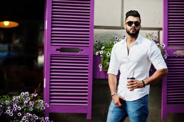 Şık uzun Arap adam modelinde beyaz gömlek, kot pantolon ve güneş gözlüğü şehir Caddesi'nde poz verdi. Kahve mor windows karşı sakal çekici Arap adamla.