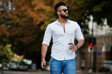 Şık uzun Arap adam modelinde beyaz gömlek, kot pantolon ve güneş gözlüğü şehir Caddesi'nde poz verdi. Fincan kahve yürüme çekici Arap adam sakal.