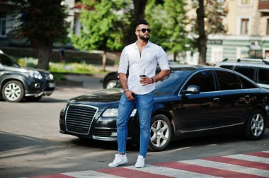 Şık uzun Arap adam modelinde beyaz gömlek, kot pantolon ve güneş gözlüğü şehir Caddesi'nde poz verdi. Kahve siyah araba karşı sakal çekici Arap adamla.