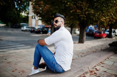 Şık uzun Arap adam modelinde beyaz gömlek, kot pantolon ve şehir Caddesi'nde oturan güneş gözlüğü. Sakal çekici Arap adam Park.