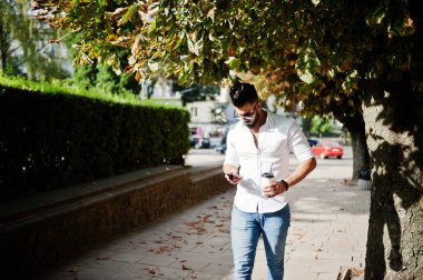 Şık uzun Arap adam modelinde beyaz gömlek, kot pantolon ve şehir Caddesi'nde yürürken güneş gözlüğü. Fincan kahve ve cep telefonu elinde sakal çekici Arap adamla.