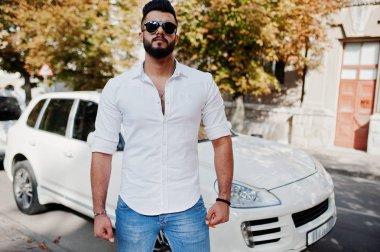 Şık uzun Arap adam modelinde beyaz gömlek, kot pantolon ve güneş gözlüğü şehir Caddesi'nde poz verdi. Sakal zengin çekici Arap adam beyaz jip araba karşı.