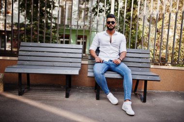Şık uzun Arap adam modelinde beyaz gömlek, kot pantolon ve güneş gözlüğü şehir Caddesi'nde poz verdi. Sakal çekici Arap adam bankta oturan.
