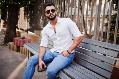 Şık uzun Arap adam modelinde beyaz gömlek, kot pantolon ve güneş gözlüğü şehir Caddesi'nde poz verdi. Sakal çekici Arap adam bankta oturan.