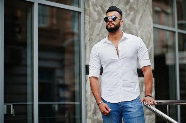 Stylish tall arabian man model in white shirt, jeans and sunglasses posed at street of city. Beard attractive arab guy.