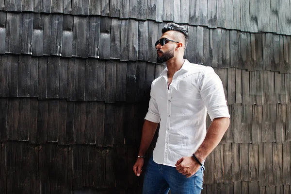 Stylish tall arabian man model in white shirt, jeans and sunglasses posed against wooden wall indoor. Beard attractive arab guy.