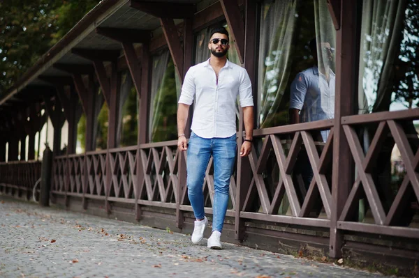 Şık uzun Arap adam modelinde beyaz gömlek, kot pantolon ve şehir Caddesi'nde yürürken güneş gözlüğü. Sakal çekici Arap adam.