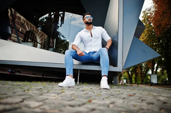 Şık uzun Arap adam modelinde beyaz gömlek, kot pantolon ve şehir Caddesi'nde oturan güneş gözlüğü. Sakal çekici Arap adam.