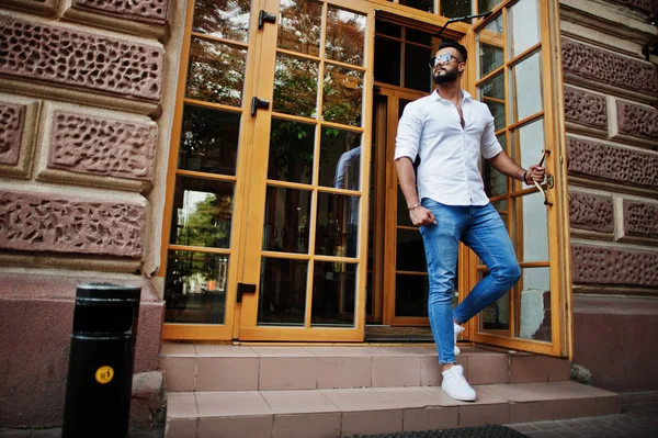 Stylish tall arabian man model in white shirt, jeans and sunglasses posed at street of city. Beard attractive arab guy against entrance door of restaurant.