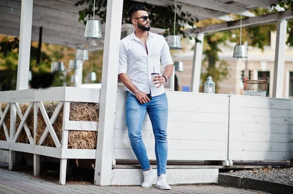 Stylish tall arabian man model in white shirt, jeans and sunglasses posed at street of city. Beard attractive arab guy with cup of coffee against outdoor cafe.