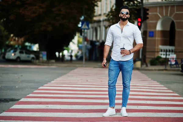 Stylish tall arabian man model in white shirt, jeans and sunglasses posed at street of city. Beard attractive arab guy with cup of coffee walking on pedestrian crossing or crosswalk.