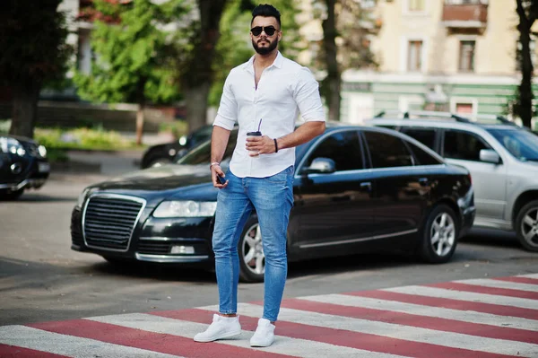 Şık uzun Arap adam modelinde beyaz gömlek, kot pantolon ve güneş gözlüğü şehir Caddesi'nde poz verdi. Kahve siyah araba karşı sakal çekici Arap adamla.