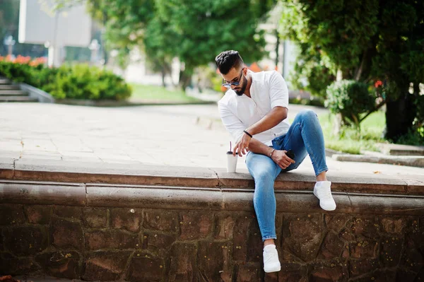 Şık uzun Arap adam modelinde beyaz gömlek, kot pantolon ve şehir Caddesi'nde oturan güneş gözlüğü. Kahve Park'ta sakal çekici Arap adamla.