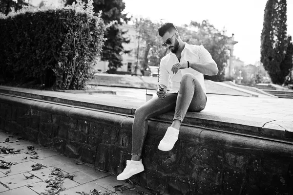 Şık uzun Arap adam modelinde beyaz gömlek, kot pantolon ve şehir Caddesi'nde oturan güneş gözlüğü. Kahve Park'ta sakal çekici Arap adamla.