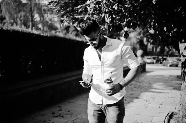 Şık uzun Arap adam modelinde beyaz gömlek, kot pantolon ve şehir Caddesi'nde yürürken güneş gözlüğü. Fincan kahve ve cep telefonu elinde sakal çekici Arap adamla.