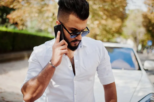 Stylish tall arabian man model in white shirt, jeans and sunglasses posed at street of city. Beard rich attractive arab guy against white suv car, speaking on mobile phone.