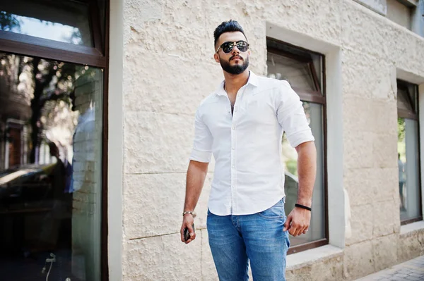 Stylish tall arabian man model in white shirt, jeans and sunglasses posed at street of city. Beard attractive arab guy.