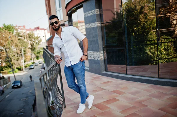 Şık uzun Arap adam modelinde beyaz gömlek, kot pantolon ve güneş gözlüğü şehir Caddesi'nde poz verdi. Sakal çekici Arap adam modern bina karşı.