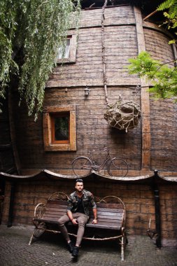 Ahşap ev karşı bankta oturmuş müthiş güzel uzun boylu ararbian sakal maço erkek gözlük ve askeri ceket açık, poz verdi.