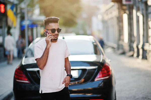 Stylish indian beard man at sunglasses and pink t-shirt against luxury car and speaking on mobile phone. India rich model posed outdoor at streets of city.