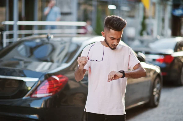 Stylish indian beard man at sunglasses and pink t-shirt against luxury car. India rich model posed outdoor at streets of city and looking at his watches.