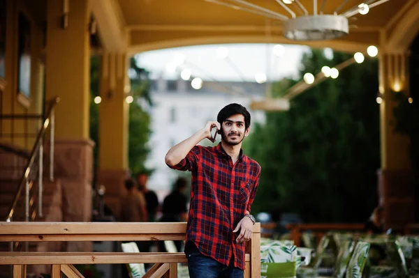 Young indian student man at red checkered shirt and jeans posed against evening cafe and speaking on phone.