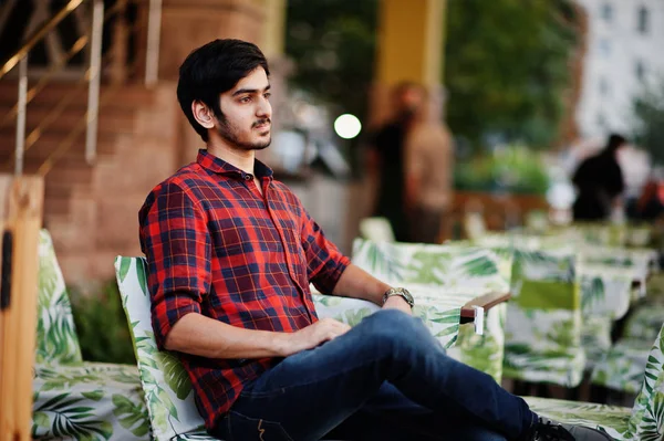 Young indian student man at red checkered shirt and jeans sitting at evening cafe.