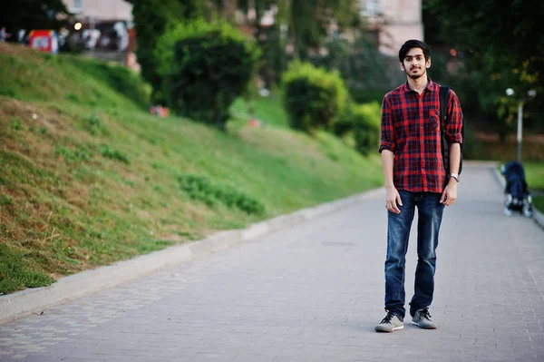 Young indian student man at red checkered shirt and jeans with backpack posed at street.