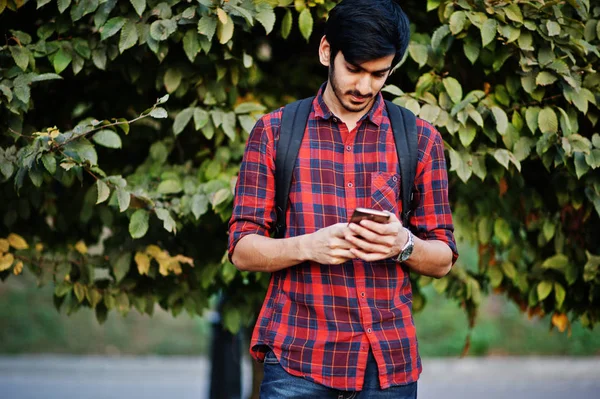Young indian student man at red checkered shirt and jeans with backpack posed at street and looking on mobile phone.