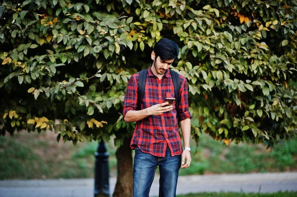 Young indian student man at red checkered shirt and jeans with backpack posed at street and looking on mobile phone.