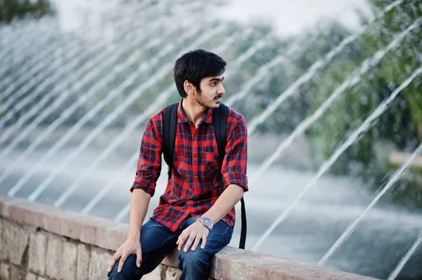 Young indian student man at checkered shirt and jeans with backpack posed on evening city against fountains.