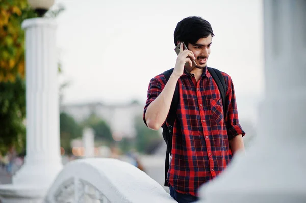 Young indian student man at checkered shirt and jeans with backpack speaking on mobile phone.