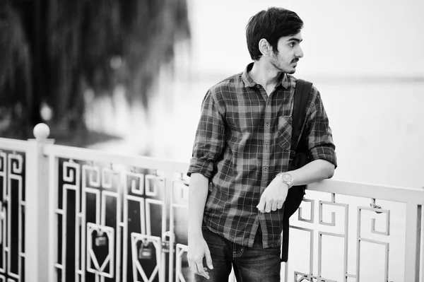 Young indian student man at checkered shirt and jeans with backpack posed on evening city against lake.