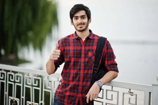 Young indian student man at checkered shirt and jeans with backpack posed on evening city against lake, shows thumb up.